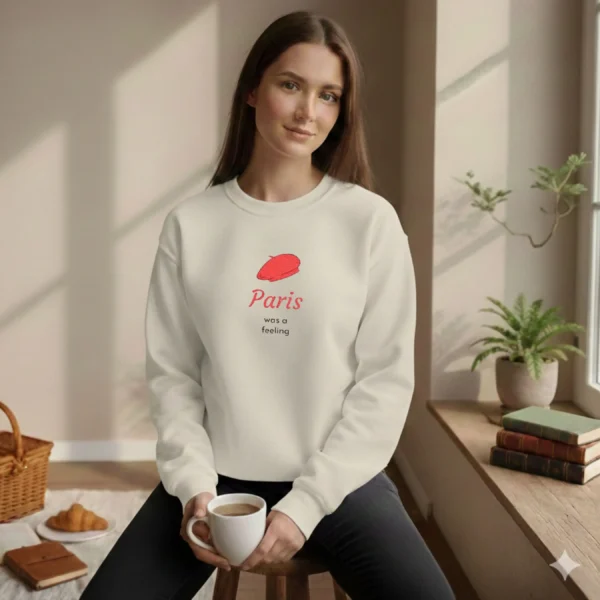 Model wearing a ash sweatshirt with a red beret design sitting by a sunny window with coffee and a croissant.