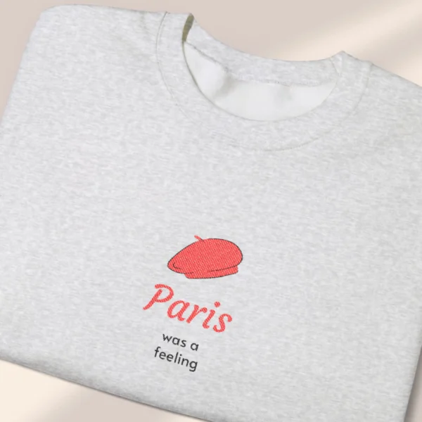 Detailed close-up of a neatly folded ash sweatshirt featuring a minimalist red beret Paris graphic.