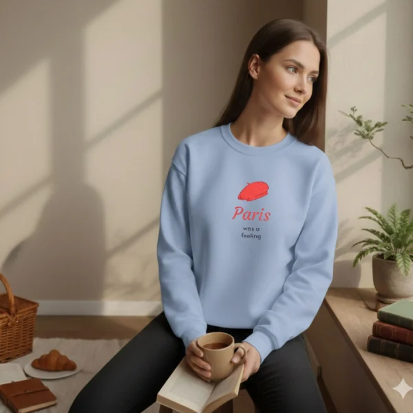 Model wearing a light blue sweatshirt with a red beret design sitting by a sunny window with coffee and a croissant.