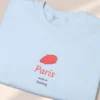 Detailed close-up of a neatly folded light blue sweatshirt featuring a minimalist red beret Paris graphic.