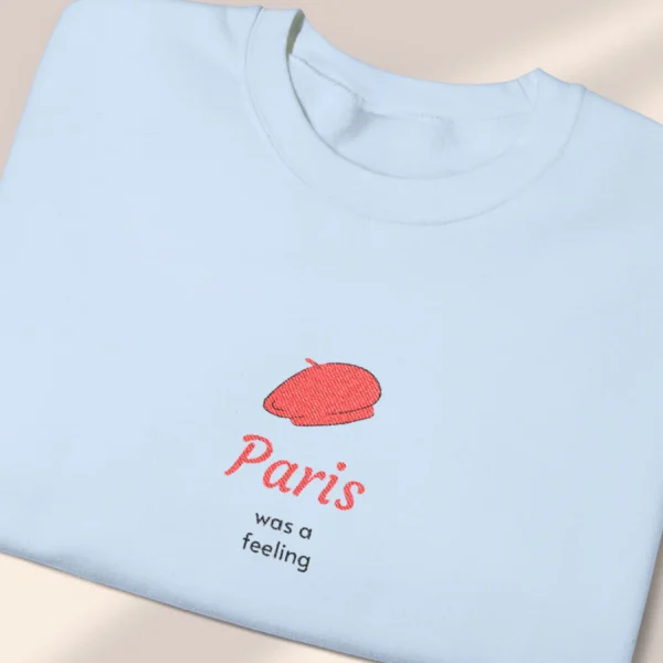 Detailed close-up of a neatly folded light blue sweatshirt featuring a minimalist red beret Paris graphic.