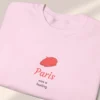 Detailed close-up of a neatly folded light pink sweatshirt featuring a minimalist red beret Paris graphic.