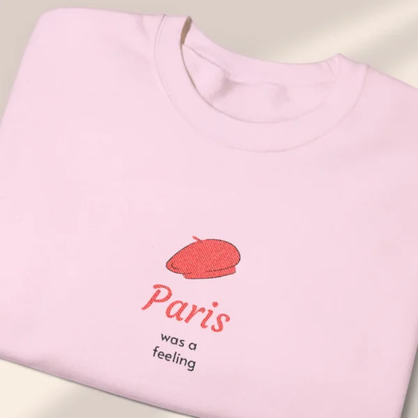 Detailed close-up of a neatly folded light pink sweatshirt featuring a minimalist red beret Paris graphic.