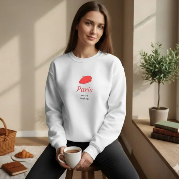 Model wearing a White sweatshirt with a red beret design sitting by a sunny window with coffee and a croissant.