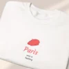 Detailed close-up of a neatly folded white sweatshirt featuring a minimalist red beret Paris graphic.