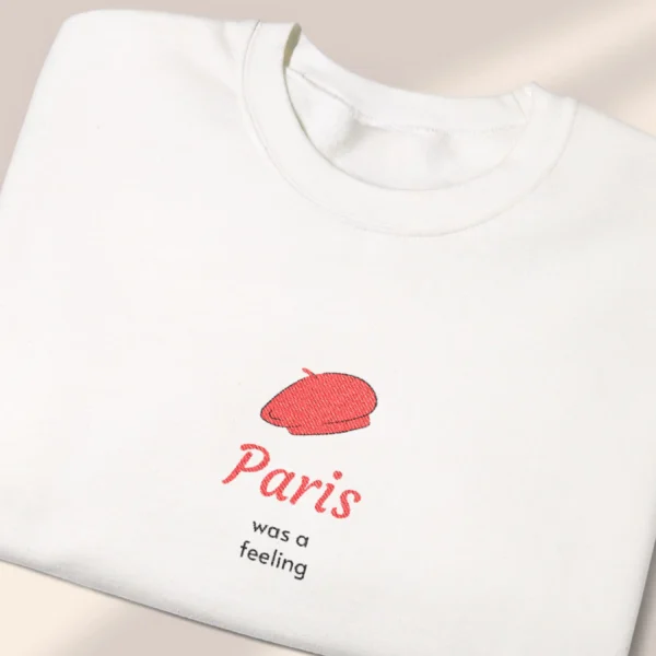 Detailed close-up of a neatly folded white sweatshirt featuring a minimalist red beret Paris graphic.